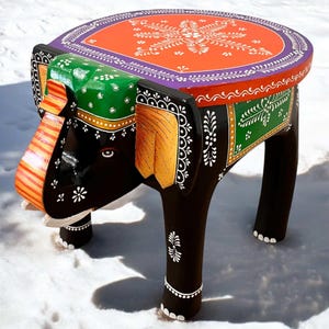 May include: Hand-painted wooden elephant stool with a colorful round top. The elephant is black with white accents and has green, orange, and purple details. The top is orange with a white floral design and a purple border.