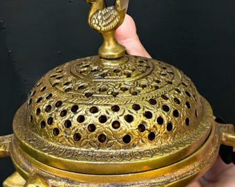 May include: A brass incense burner with a turtle-shaped base and a decorative lid featuring a floral pattern and small holes. A peacock figurine adorns the top. The burner is held in a hand against a dark background.