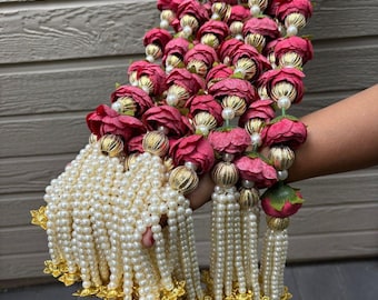 May include: A decorative garland featuring burgundy-colored fabric roses, gold accents, and strands of faux pearls. The garland has multiple tassels of pearls with gold-colored decorative elements at the base.