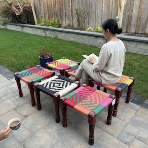 May include: Four square stools with dark brown wooden legs and woven seats featuring colorful geometric patterns in shades of pink, green, orange, yellow, blue, black, and white, arranged on a stone patio surface outdoors.