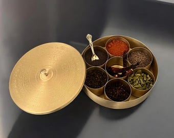 Op de afbeelding: Een goudkleurige kruidenbox met een decoratief deksel en een kleine lepel. De box is verdeeld in verschillende compartimenten, elk gevuld met verschillende kruiden, waaronder zwarte peperkorrels, rode chilipepers en groene kardemompeulen.