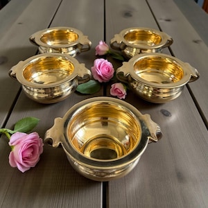 May include: Five small, gold-coloured bowls with ornate handles are arranged on a wooden surface. Pink roses are scattered around the bowls. The bowls have a shiny, reflective surface, adding a touch of colour.