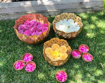Puede incluir: Tres cuencos decorativos de metal dorado llenos de flores. Uno contiene rosas moradas, otro blancas y el tercero amarillas. También hay cuencos más pequeños con pétalos rosas, todo sobre un césped verde.