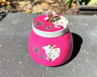 May include: A vibrant pink jar with a decorative lid. The lid features a crystal knob and a design with palm trees and a cow illustration. The jar's body also has a cow illustration and palm tree accents.