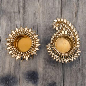 May include: Two decorative gold candle holders. One is round with a sunburst design, and the other is paisley-shaped. Both have a central well for a candle and are embellished with small mirrors and clear crystals.