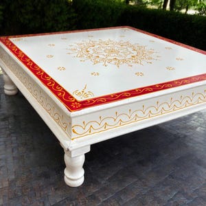 Peut inclure: Table basse carrée blanche avec des détails peints en rouge et or. Le plateau de la table présente un grand motif de mandala avec une bordure rouge. La table a quatre pieds blancs.