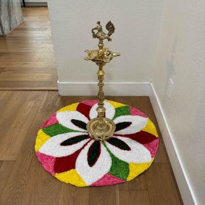 Bright rangoli mat featuring oversized white petals with colorful border, topped with a traditional brass lamp, placed indoors near a wall