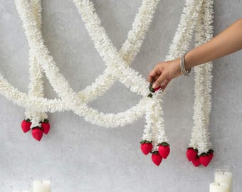 May include: A long white floral garland featuring multiple strands of small white petals. The garland is accented with small, bright red fabric strawberry charms attached at the ends and intervals. Two white pillar candles sit on the surface.