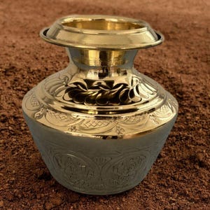 May include: A polished brass pot featuring a wide, flared rim and a short, cylindrical neck. The body of the vessel is decorated with intricate, embossed floral patterns and geometric motifs, showcasing a reflective, metallic gold finish.