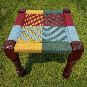 May include: A square stool with four dark brown wooden legs and a woven seat. The seat features a geometric pattern created with thick cord in shades of yellow, light blue, dark green, and deep red, arranged in a repeating chevron design.