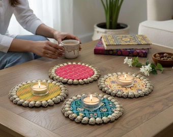 Puede incluir: Cuatro posavasos circulares de tela con bordes de cuentas metálicas, cada uno con un patrón colorido único. Tres posavasos sostienen portavelas de vidrio transparente con velas encendidas. Los posavasos están sobre una superficie de madera.