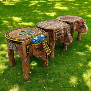 May include: Three wooden elephant stools with intricately painted designs. The stools have brown bodies, with colorful accents in blue, yellow, and white. The tops of the stools feature floral and geometric patterns. The stools are placed on a green grassy surface.
