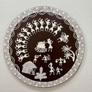 May include: A round, brown wooden plate with a white design depicting a group of people dancing in a circle around a drum. The design also includes a hut, a tree, and other figures.
