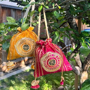 Peut inclure: Deux sacs à cordon décoratifs, l'un jaune doré et l'autre fuchsia, suspendus à une branche d'arbre. Chaque sac présente un motif circulaire brodé avec une bordure dorée et une sangle perlée. Des glands pendent du bas de chaque sac.
