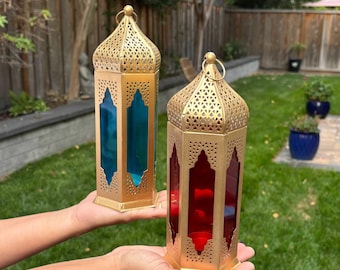 May include: Two gold-colored metal lanterns with intricate cut-out designs. One lantern has blue glass panels, the other red. Both have a ring at the top for hanging. The lanterns are held up in front of a green lawn.