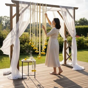 Woman arranging decorations on a wooden arch with white curtains and yellow hanging elements in a garden setting.