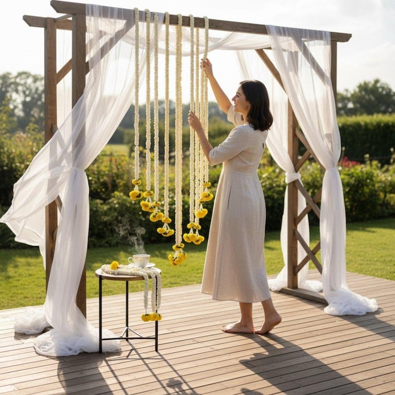 Woman arranging decorations on a wooden arch with white curtains and yellow hanging elements in a garden setting.