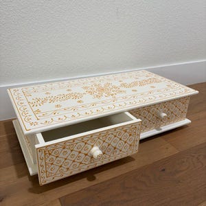 May include: A white wooden jewellery box with two drawers, decorated with gold floral patterns. The top and drawer fronts feature intricate designs, and the drawers have round, white knobs. The box is open, revealing an empty drawer.