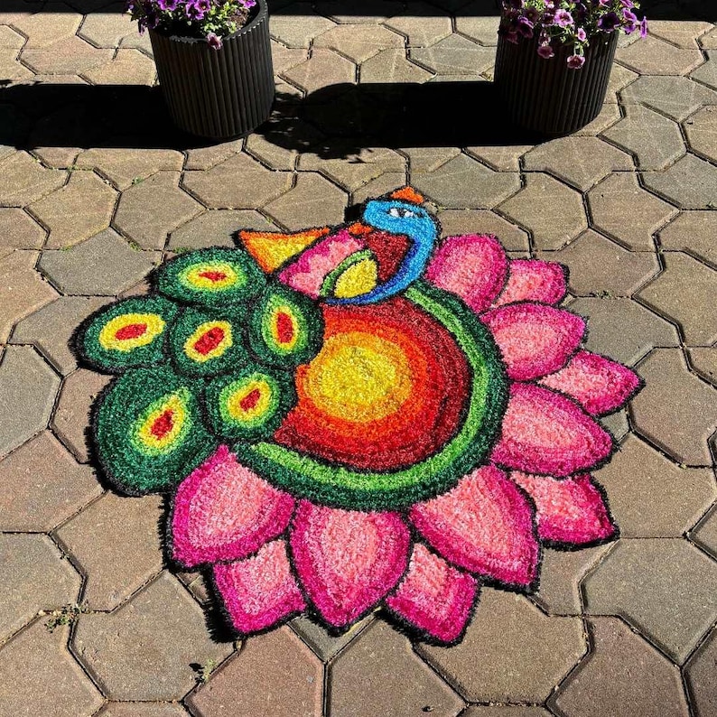 Vibrant peacock-themed rangoli mat with green outer petals and a stylized bird motif at the center, displayed on stone flooring