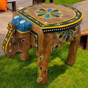 May include: A handcrafted wooden elephant-shaped side table. The table features a brown finish with intricate hand-painted details in blue, yellow, and white. The top has a floral design. The table is on a green lawn.