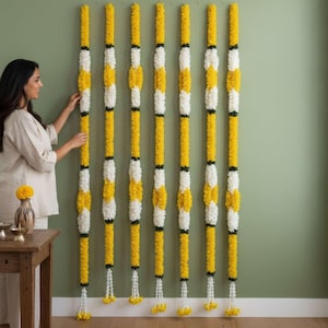May include: Seven vertical floral garlands hanging against a wall, each featuring alternating segments of bright yellow and white marigold flowers. The garlands are long and slender, with small white tassels at the bottom of each individual strand.