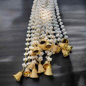 May include: A white garland with small white flowers and gold bells hanging from the bottom. The garland is hanging on a dark gray background.