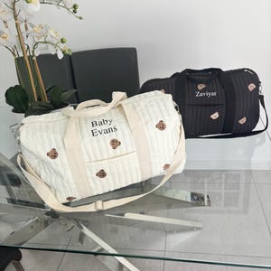 May include: Two quilted duffel bags, one white and one black, featuring embroidered teddy bear motifs and personalized text. The white bag displays the name Baby Evans, while the black bag displays the name Zaviyar. Both bags have handles.