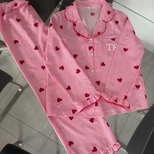 May include: Pink and white striped pajama set with red heart accents. The long-sleeved top has a scalloped collar and cuffs trimmed in red. The pants have a ruffled hem. The initials "TF" are embroidered on the left chest.