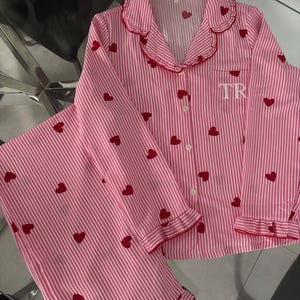 May include: Pink and white striped pajama set with red heart accents. The long-sleeved top has a ruffled collar and cuffs, with the initials "TR" embroidered on the chest. The pajama pants have a ruffled hem.