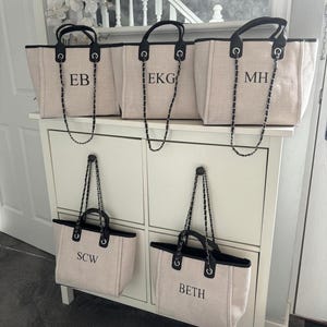 May include: Five beige canvas tote bags with black trim and silver chain straps. Each bag has a different name monogrammed on the front: EB, EKG, MH, SCW, and BETH.