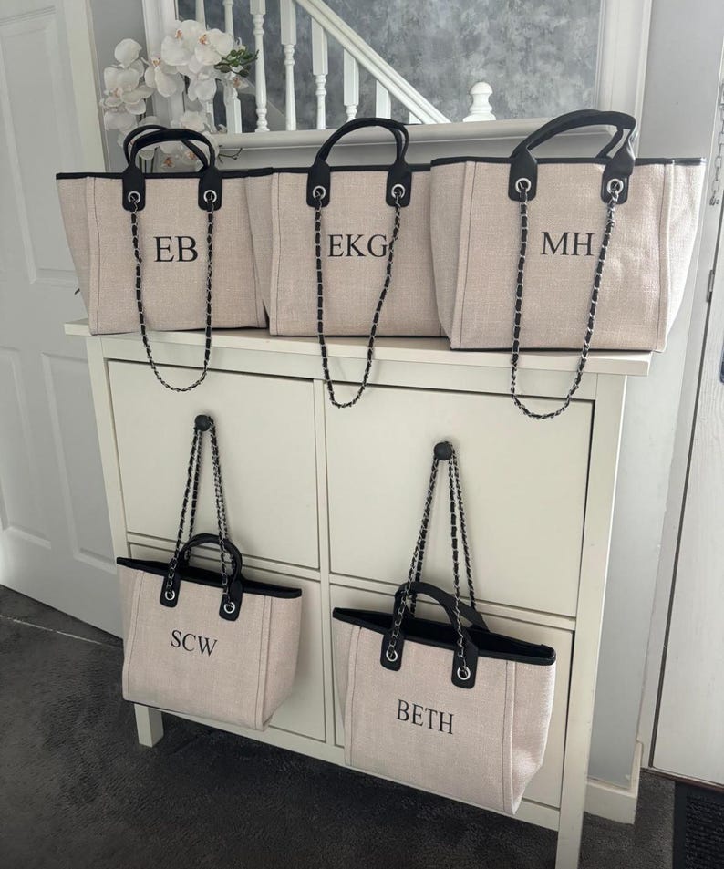 May include: Five beige canvas tote bags with black trim and silver chain straps. Each bag has a different name monogrammed on the front: EB, EKG, MH, SCW, and BETH.