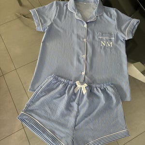 May include: Blue and white striped pajama set. The short-sleeved top has a breast pocket with the initials "NM" embroidered in white. The shorts have a white bow and trim. The set is made of a lightweight fabric.