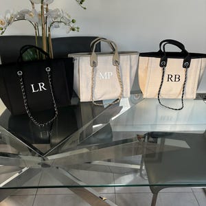 May include: Three canvas tote bags with chain straps. The bags are personalized with the initials "LR", "MP", and "RB". The bags are on a glass table with a silver metal base.