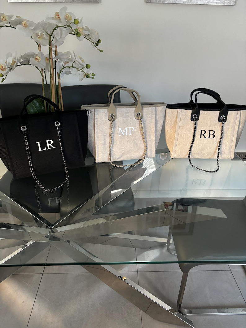 May include: Three canvas tote bags with chain straps. The bags are personalized with the initials "LR", "MP", and "RB". The bags are on a glass table with a silver metal base.