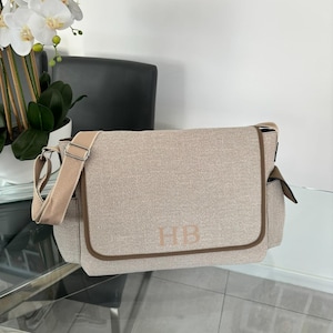 May include: A beige canvas diaper bag with a brown trim and a brown leather strap. The bag has the initials "HB" embroidered on the front.