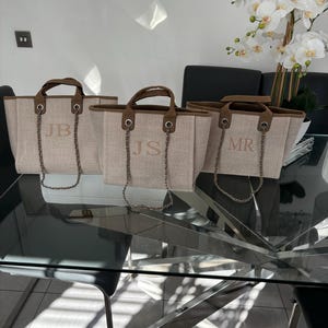 May include: Three beige canvas tote bags with brown leather handles and silver chain straps. The bags have the initials "JB", "JS", and "MR" embroidered on the front.