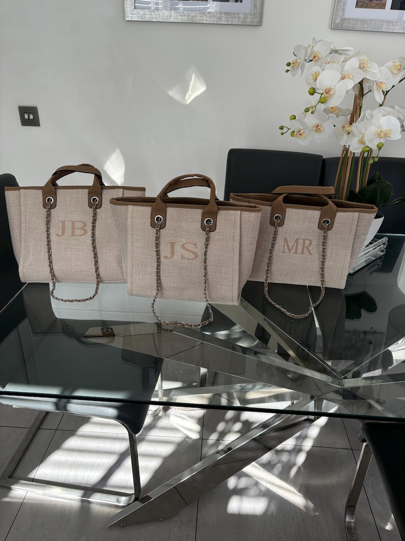May include: Three beige canvas tote bags with brown leather handles and silver chain straps. The bags have the initials "JB", "JS", and "MR" embroidered on the front.