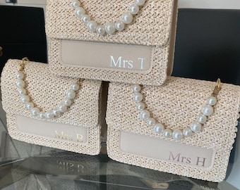 May include: Three rectangular straw handbags with a woven texture, featuring a flap closure and a handle made of large white faux pearls. Each bag has a smooth beige panel on the front with personalized text reading Mrs T, Mrs B, or Mrs H.
