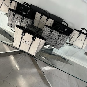 May include: A group of seven white canvas tote bags with black trim and black chain straps. Each bag has a different set of black embroidered letters on the front. The bags are arranged on a glass table with a silver metal base.