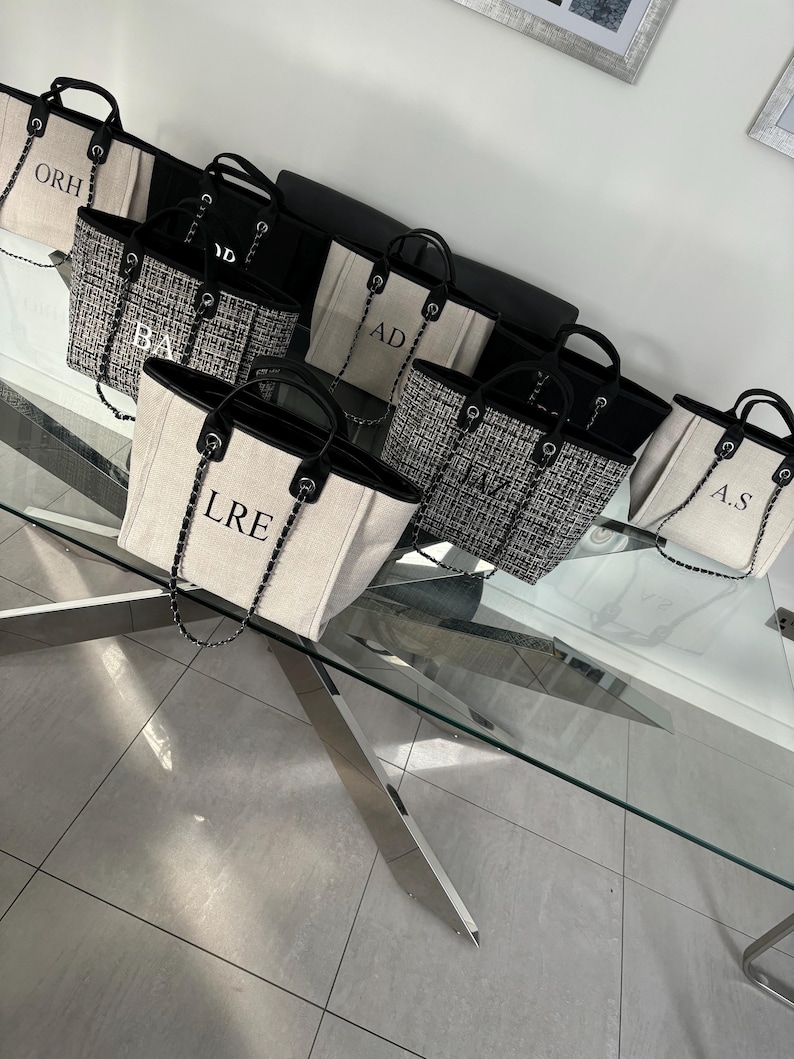 May include: A group of seven white canvas tote bags with black trim and black chain straps. Each bag has a different set of black embroidered letters on the front. The bags are arranged on a glass table with a silver metal base.