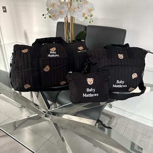 May include: A set of four black quilted baby bags with teddy bear appliqués. The bags are different sizes, including a tote, a diaper bag, and a small pouch. The text "Baby Matthews" is printed on the bags.