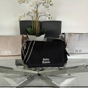 May include: Three different diaper bags with personalized embroidery. The bags are beige, black, and gray. The beige bag has the letters "ORW" embroidered on it. The black bag has the words "Baby Renzo" embroidered on it. The gray bag has the letters "B.B." embroidered on it.