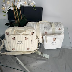 May include: Cream-colored baby bags with bear embroidery. The bags include a diaper bag, a tote bag, and a small pouch. The text "Baby Tavares" is printed on the bags. The bags are on a glass table with a white flower arrangement in the background.