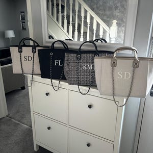 May include: Four canvas tote bags with black chain straps and personalised initials. The bags are lined up on a white dresser with black drawer pulls. The initials on the bags are "SD", "FL", "KM", and "SD".
