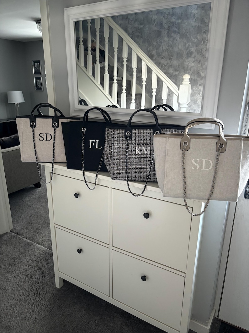 May include: Four canvas tote bags with black chain straps and personalized initials. The bags are lined up on a white dresser with black drawer pulls. The initials on the bags are "SD", "FL", "KM", and "SD".