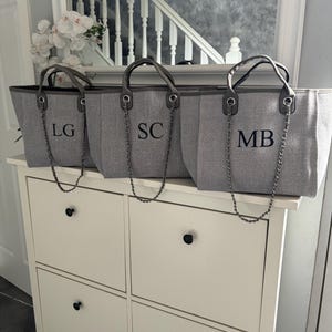 May include: Three gray canvas tote bags with leather handles and silver chain straps. The bags are monogrammed with the initials "LG", "SC", and "MB".