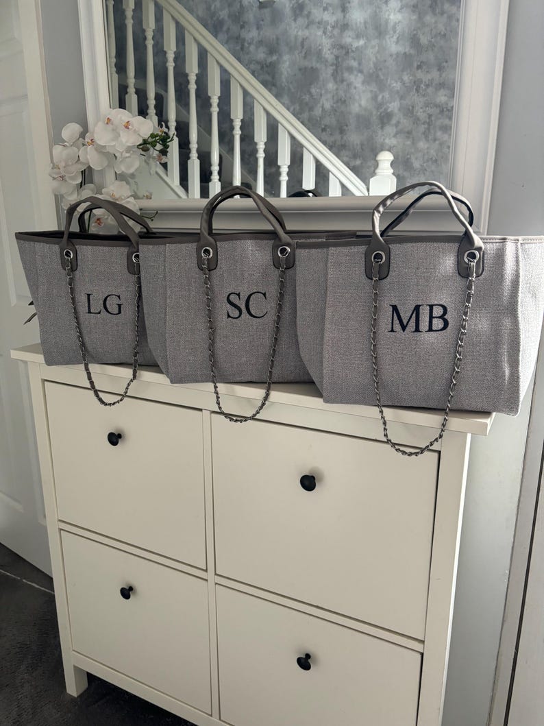 May include: Three gray canvas tote bags with leather handles and silver chain straps. The bags are monogrammed with the initials "LG", "SC", and "MB".