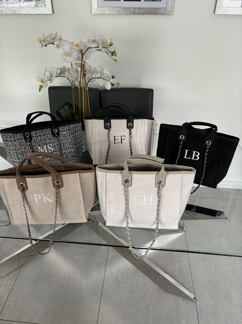 May include: Five tote bags with different initials embroidered on them. The bags are made of a beige canvas material with a brown leather handle and a silver chain strap. The initials are "MS", "EF", "LB", "PK", and "EH".