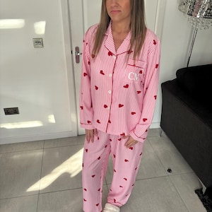May include: Pink and white striped pajama set with red heart accents. The long-sleeved top has a breast pocket with embroidered initials. The matching pants and top are button-down. The person is wearing white fuzzy slippers.