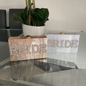 May include: Two rectangular box clutches with a marbled finish, one in beige and one in white. Each clutch features the word BRIDE in large, raised, rhinestone-encrusted letters on the front, with a gold-toned metal clasp closure on the top edge.